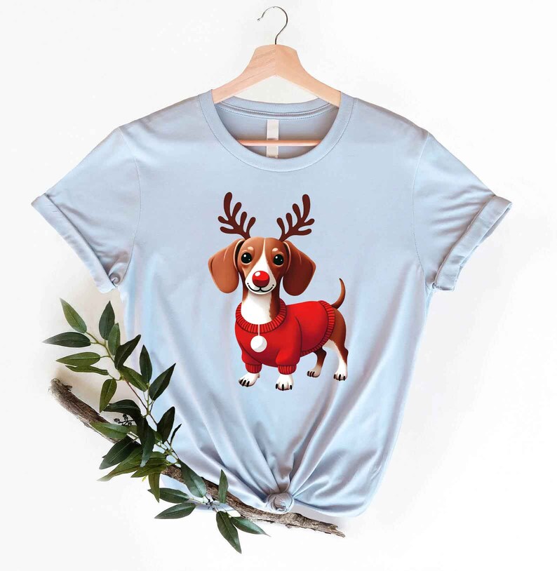 May include: A light blue t-shirt with a cartoon image of a dachshund wearing a red sweater and reindeer antlers. The dachshund has a red nose and white markings on its face.