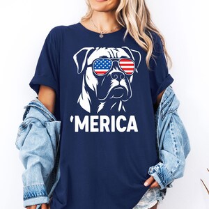 May include: Dark blue t-shirt with a white graphic of a dog wearing sunglasses in the style of the American flag. The word "'MERICA" is printed below the dog illustration. The shirt has short sleeves and a casual fit.