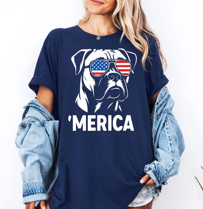 May include: Dark blue t-shirt with a white graphic of a dog wearing sunglasses in the style of the American flag. The word "'MERICA" is printed below the dog illustration. The shirt has short sleeves and a casual fit.