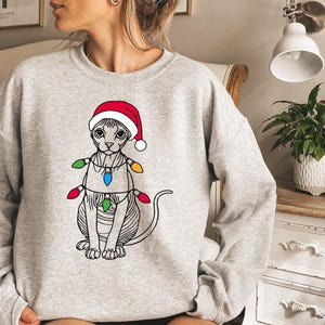 May include: A gray sweatshirt with a graphic of a cat wearing a Santa hat and Christmas lights. The cat is a Sphynx breed with no fur.