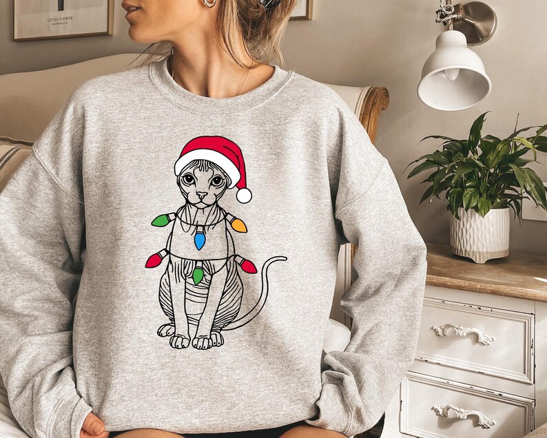 May include: A gray sweatshirt with a graphic of a cat wearing a Santa hat and Christmas lights. The cat is a Sphynx breed with no fur.