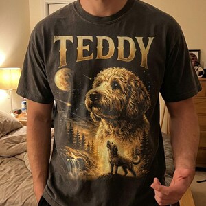 May include: Dark gray t-shirt featuring a detailed illustration of a dog with the name "TEDDY" in gold lettering. The design includes a night sky, moon, waterfall, and trees, creating a scenic landscape.