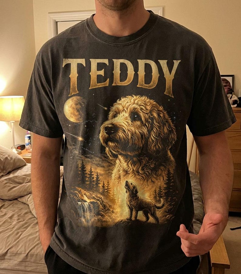 May include: Dark gray t-shirt featuring a detailed illustration of a dog with the name "TEDDY" in gold lettering. The design includes a night sky, moon, waterfall, and trees, creating a scenic landscape.