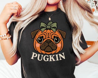 Könnte beinhalten: Schwarzes T-Shirt mit einem Cartoon-Mops, der als Kürbis mit einer grünen Schleife gestaltet ist. Das Wort "PUGKIN" ist weiß unter der Grafik aufgedruckt. Das Shirt ist ein bequemer, lässiger Stil.