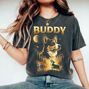 May include: Dark gray t-shirt featuring a detailed illustration of a corgi dog with the word "BUDDY" in gold lettering. The design includes a moon, forest, and a smaller corgi. The shirt is a vintage style.