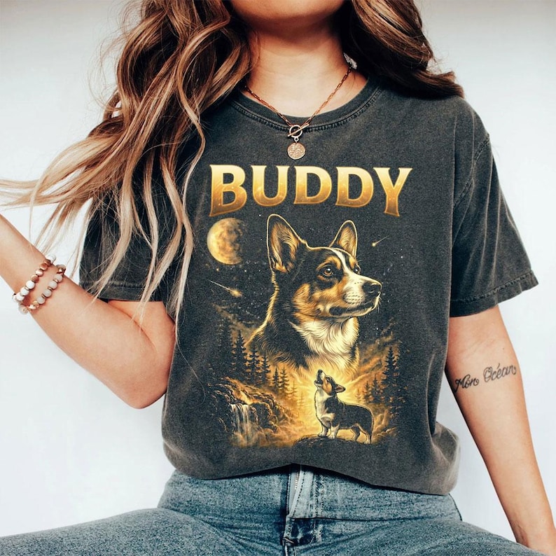 May include: Dark gray t-shirt featuring a detailed illustration of a corgi dog with the word "BUDDY" in gold lettering. The design includes a moon, forest, and a smaller corgi. The shirt is a vintage style.