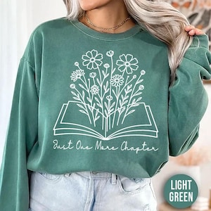 Op de afbeelding: Lichtgroene sweatshirt met een wit bloemenontwerp dat uit een open boek komt. De tekst "Just One More Chapter" staat onder het boek. De woorden "LIGHT GREEN" staan in een kader rechtsonder. De sweatshirt heeft een casual stijl.