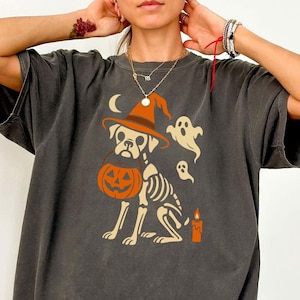 Könnte beinhalten: Dunkelgraues T-Shirt mit einem Halloween-Motiv. Das Design zeigt einen Hund mit Hexenhut und Kürbis-Eimer, mit Skelett-Details. Zusätzliche Elemente sind Geister, ein Halbmond und eine Kerze.