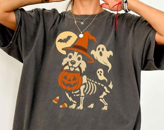 May include: Dark gray t-shirt featuring a Halloween-themed graphic. The design includes a dog wearing a witch hat and holding a jack-o'-lantern, with a skeleton body, ghosts, a bat, and candy corn.