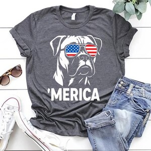 May include: Dark grey t-shirt with a white graphic of a dog wearing sunglasses in the style of the American flag. The word "'MERICA" is printed below. The shirt is shown with blue jeans and white trainers.