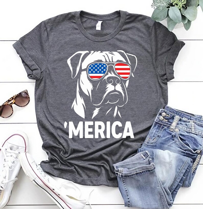 May include: Dark grey t-shirt with a white graphic of a dog wearing sunglasses in the style of the American flag. The word "'MERICA" is printed below. The shirt is shown with blue jeans and white trainers.