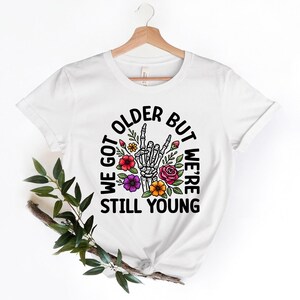 May include: White t-shirt with a black circle graphic featuring a skeleton hand giving a rock and roll sign, surrounded by colorful flowers. The text reads "We got older but we're still young".