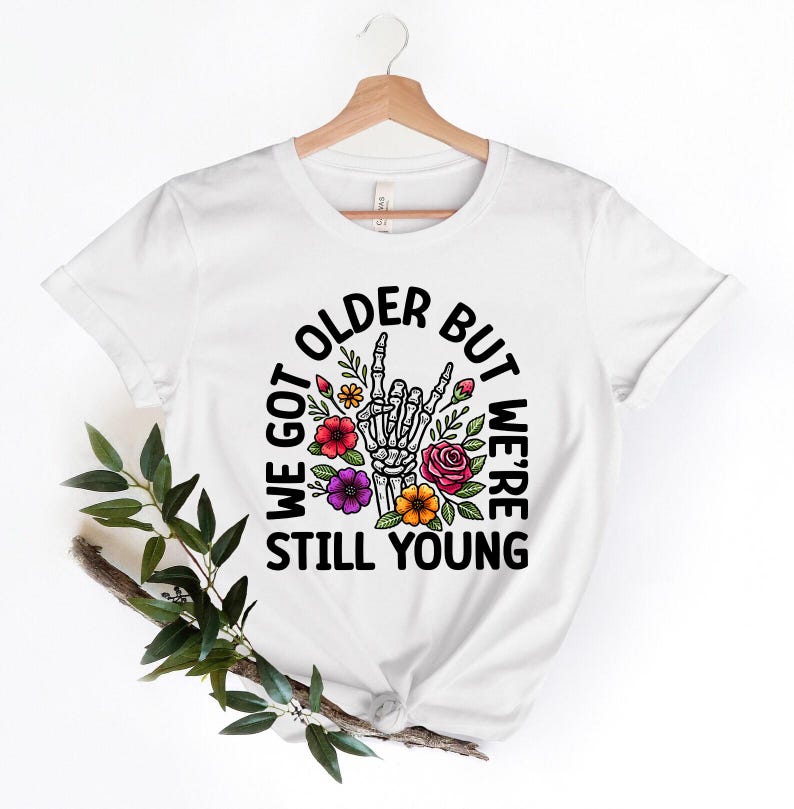 May include: White t-shirt with a black circle graphic featuring a skeleton hand giving a rock and roll sign, surrounded by colorful flowers. The text reads "We got older but we're still young".