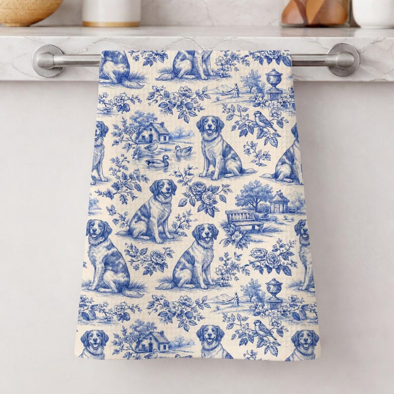 K&ouml;nnte beinhalten: Ein blau-wei&szlig; gemustertes K&uuml;chentuch mit Toile-de-Jouy-Design. Das Design zeigt Hunde, H&auml;user, Blumen und V&ouml;gel. Das Handtuch h&auml;ngt an einer silbernen Stange.