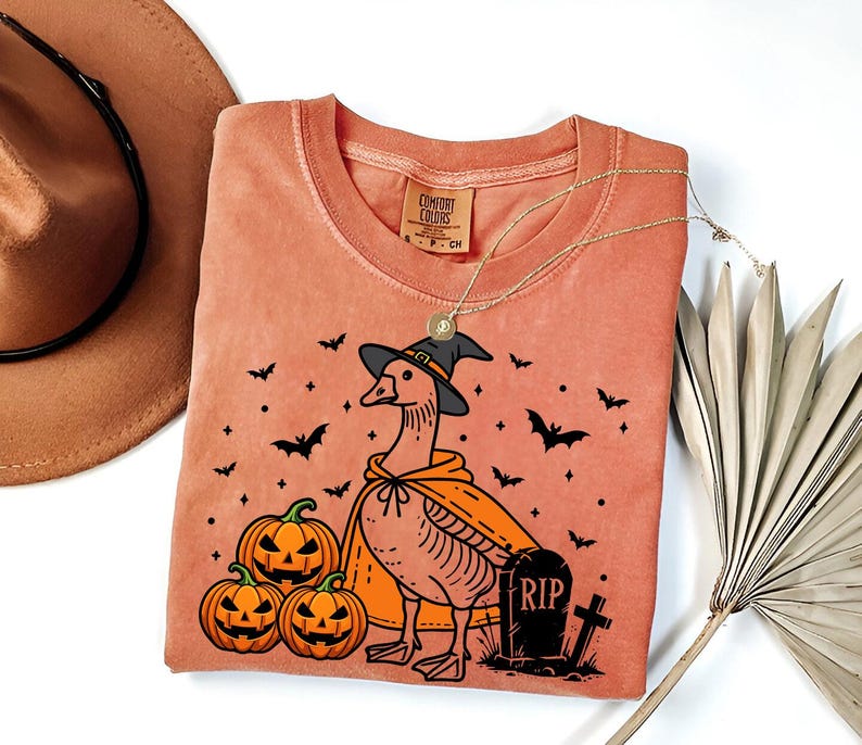 Puede incluir: Una camiseta de color melocot&oacute;n con una ilustraci&oacute;n de dibujos animados de un ganso con un sombrero de bruja y sosteniendo una calabaza. El ganso est&aacute; rodeado de murci&eacute;lagos y calabazas. El texto "RIP" est&aacute; en una l&aacute;pida.