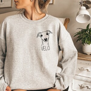K&ouml;nnte beinhalten: Ein grauer Pullover mit einer schwarzen Linienzeichnung eines Hundekopfes und dem Namen "VELO" auf der Vorderseite.
