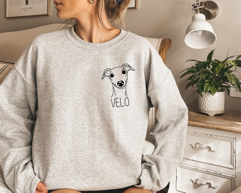 K&ouml;nnte beinhalten: Ein grauer Pullover mit einer schwarzen Linienzeichnung eines Hundekopfes und dem Namen "VELO" auf der Vorderseite.