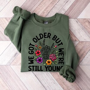 May include: Green sweatshirt with a black graphic print that says "We got older but we're still young" with a skeleton hand giving a peace sign and flowers around it.