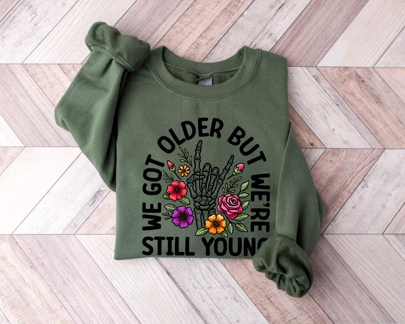 May include: Green sweatshirt with a black graphic print that says "We got older but we're still young" with a skeleton hand giving a peace sign and flowers around it.