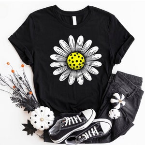 May include: Black t-shirt with a white daisy featuring a yellow pickleball in the center. The shirt is laying on a white surface with black and white pumpkins and black and white converse shoes.