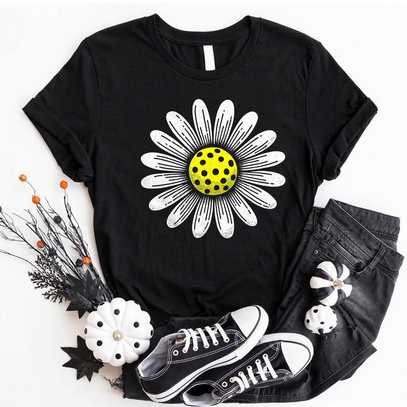 May include: Black t-shirt with a white daisy featuring a yellow pickleball in the center. The shirt is laying on a white surface with black and white pumpkins and black and white converse shoes.