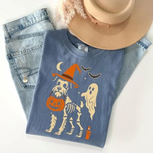 May include: A blue t-shirt featuring a Halloween-themed design. The graphic includes a dog wearing a witch hat and carrying a pumpkin, with a ghost, bats, and a candle. The shirt is paired with light-wash jeans and a tan hat.