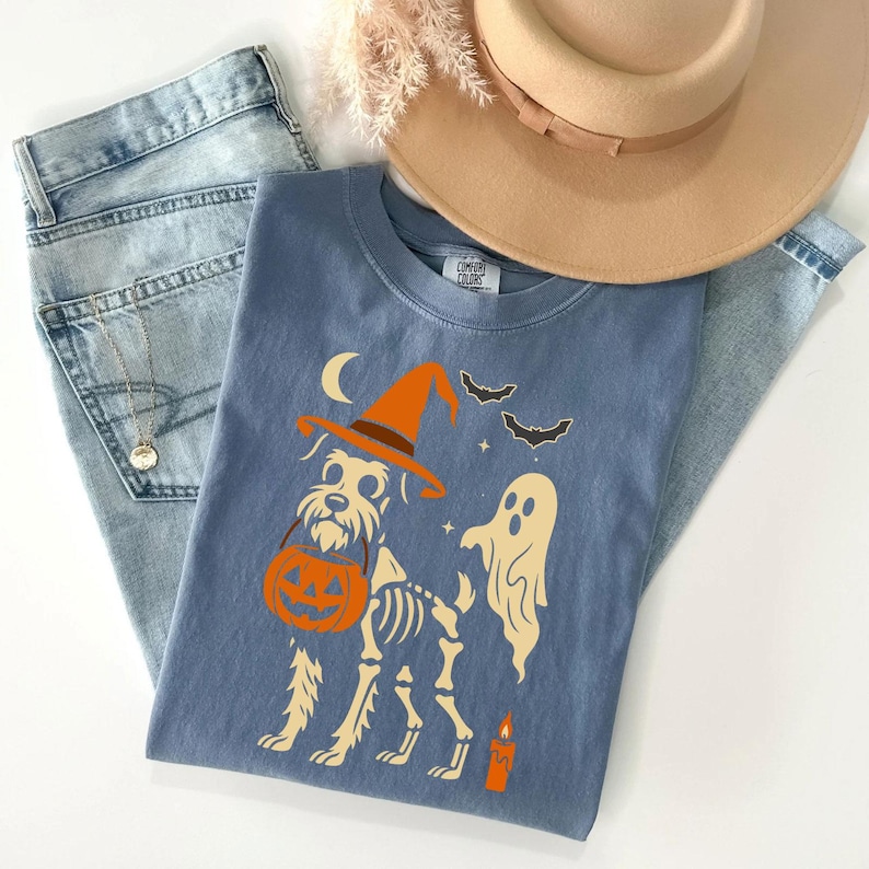 May include: A blue t-shirt featuring a Halloween-themed design. The graphic includes a dog wearing a witch hat and carrying a pumpkin, with a ghost, bats, and a candle. The shirt is paired with light-wash jeans and a tan hat.