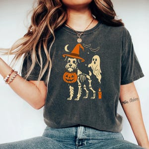 May include: Dark gray t-shirt featuring a Halloween-themed graphic. The design includes a dog wearing a witch hat and holding a jack-o'-lantern, with a skeleton, ghost, bats, moon, and candle. The shirt is a comfortable, casual style.