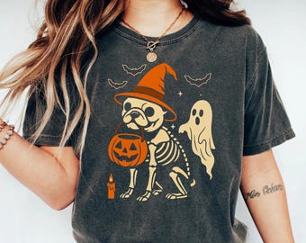 May include: Dark gray t-shirt featuring a Halloween-themed graphic. The design includes a skeleton pug wearing a witch's hat and holding a jack-o'-lantern bucket. A ghost and bats are also part of the design. A candle is lit near the dog.