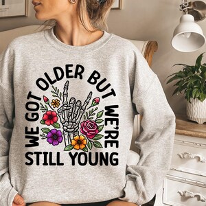 May include: Gray sweatshirt with a black graphic print that says "We got older but we're still young". The graphic also includes a skeleton hand with flowers.