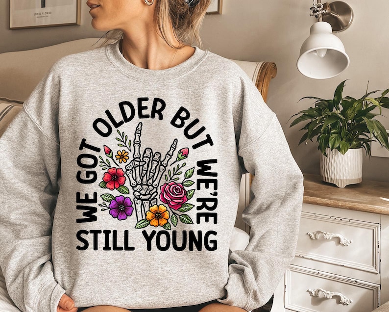 May include: Gray sweatshirt with a black graphic print that says "We got older but we're still young". The graphic also includes a skeleton hand with flowers.