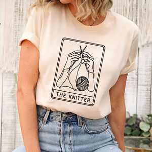 K&ouml;nnte beinhalten: Cremefarbenes T-Shirt mit einem schwarzen Strich-Design von H&auml;nden, die mit Nadeln und Garn stricken, in einem rechteckigen Rahmen. Der Text "THE KNITTER" steht darunter. Das Shirt hat hochgekrempelte &Auml;rmel.