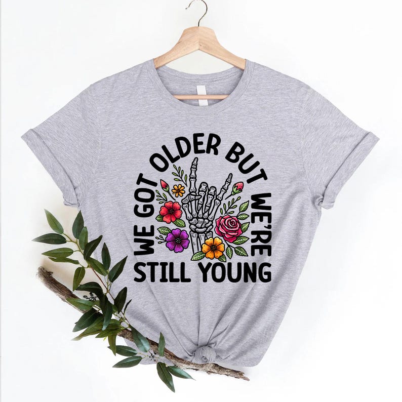 May include: Gray t-shirt with a black graphic of a skeleton hand making a rock and roll sign surrounded by flowers. The text reads "We got older but we're still young".