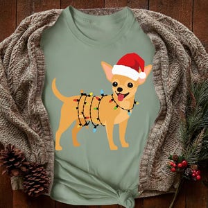 May include: A light green t-shirt featuring a cartoon chihuahua wearing a Santa hat and wrapped in Christmas lights. The dog is smiling with its tongue out. The shirt is displayed on a wooden surface with a knitted sweater.