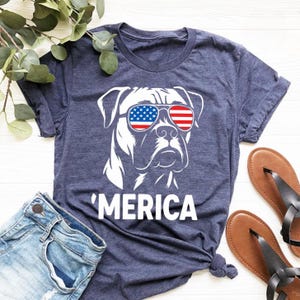 Peut inclure: T-shirt bleu marine avec un motif blanc d'un chien portant des lunettes de soleil aux couleurs du drapeau américain. Le mot "'MERICA" est imprimé en dessous. Le short en jean bleu et les sandales marron complètent l'ensemble.