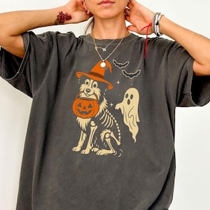May include: Dark grey t-shirt with a Halloween design. It features a dog wearing a witch's hat and carrying a jack-o'-lantern, with a skeleton outline, bats, and a ghost. The shirt has a vintage, worn appearance.