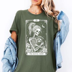 Puede incluir: Camiseta verde con un gráfico blanco de un esqueleto sosteniendo un cachorro Rottweiler. El gráfico está en el estilo de una carta de tarot y dice "The Rottie Mom" en la parte inferior.