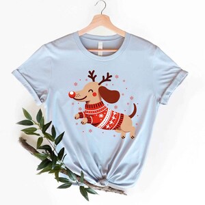 May include: Light blue t-shirt with a cartoon image of a dachshund wearing a red and white Christmas sweater and reindeer antlers. The dachshund is smiling and has a red nose.