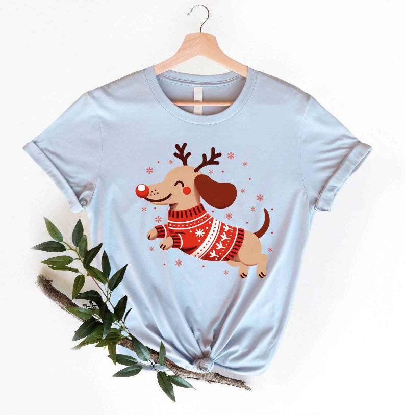 May include: Light blue t-shirt with a cartoon image of a dachshund wearing a red and white Christmas sweater and reindeer antlers. The dachshund is smiling and has a red nose.