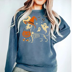 May include: A blue sweatshirt featuring a Halloween-themed graphic. The design includes a dachshund skeleton wearing a witch hat and holding a pumpkin. Additional elements include bats, ghosts, stars, and a moon.