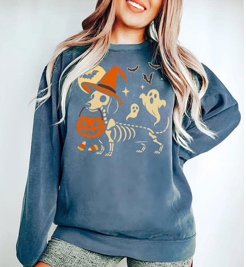 May include: A blue sweatshirt featuring a Halloween-themed graphic. The design includes a dachshund skeleton wearing a witch hat and holding a pumpkin. Additional elements include bats, ghosts, stars, and a moon.