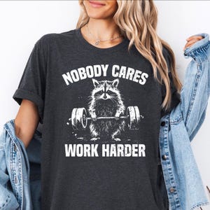 Puede incluir: Una camiseta gris oscuro con un gráfico blanco de un mapache levantando una barra de pesas. El texto "Nobody Cares Work Harder" está impreso en la camiseta.