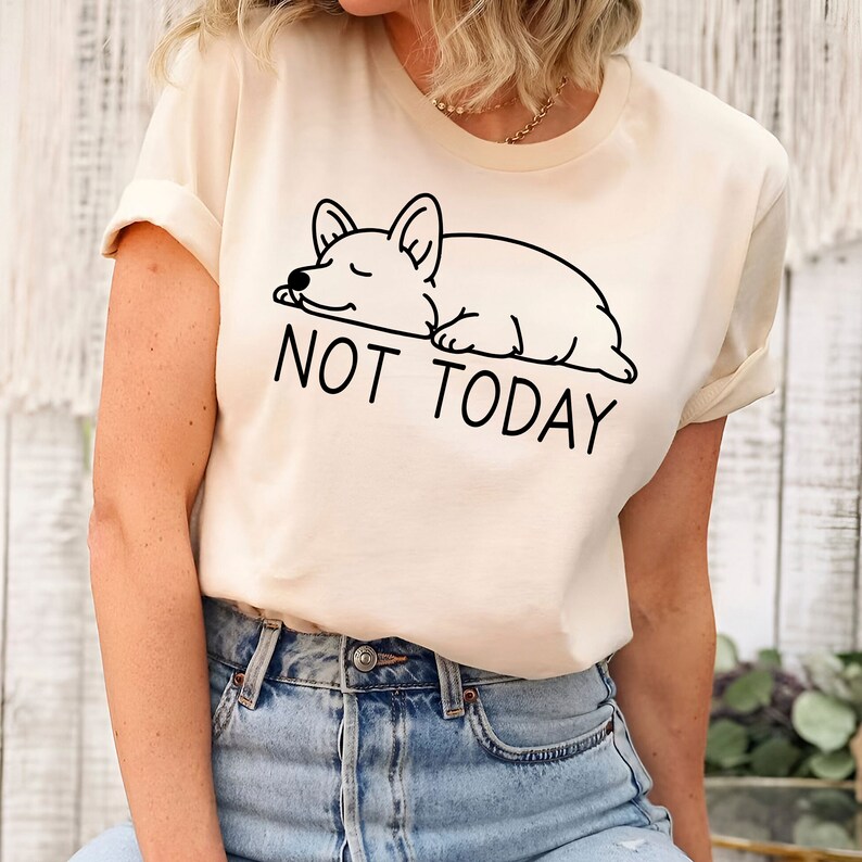 May include: A cream colored t-shirt with a black line drawing of a sleeping corgi and the text "NOT TODAY".
