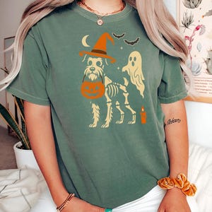 May include: Green t-shirt featuring a Halloween-themed design. The graphic includes a dog skeleton wearing a witch hat and carrying a pumpkin. A ghost, bats, a candle, and a crescent moon are also depicted.