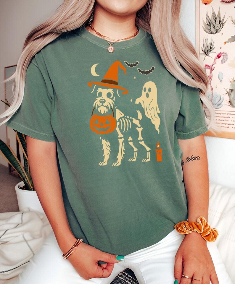 May include: Green t-shirt featuring a Halloween-themed design. The graphic includes a dog skeleton wearing a witch hat and carrying a pumpkin. A ghost, bats, a candle, and a crescent moon are also depicted.