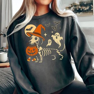 May include: Dark gray sweatshirt featuring a Halloween-themed graphic. The design includes a dachshund skeleton wearing a witch's hat and holding a jack-o'-lantern. Bats, ghosts, and a full moon complete the spooky illustration.