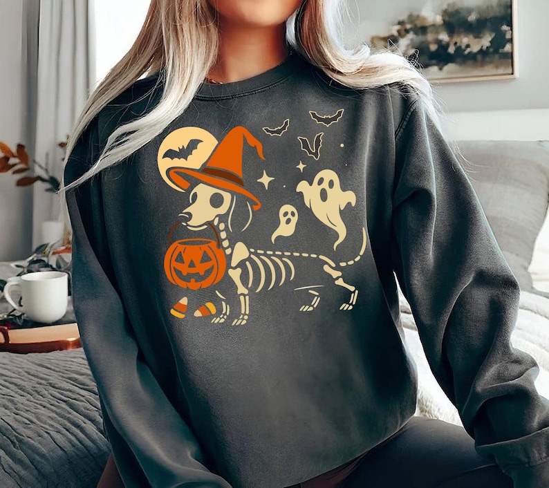 May include: Dark gray sweatshirt featuring a Halloween-themed graphic. The design includes a dachshund skeleton wearing a witch's hat and holding a jack-o'-lantern. Bats, ghosts, and a full moon complete the spooky illustration.