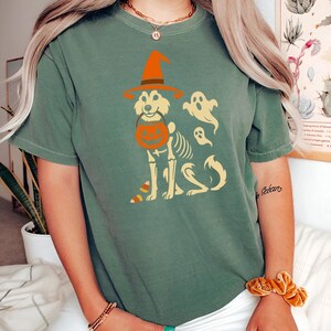 May include: Green t-shirt featuring a Halloween-themed design. The graphic includes a dog wearing a witch hat and holding a pumpkin bucket, with a skeleton body and ghost figures. The design is in orange and cream colors.