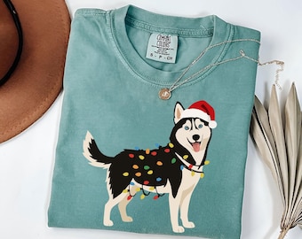 May include: A teal Comfort Colors t-shirt featuring a cartoon husky wearing a Santa hat and Christmas lights. The dog has black and white fur, blue eyes, and a red tongue. A silver necklace with a pendant is draped across the shirt.