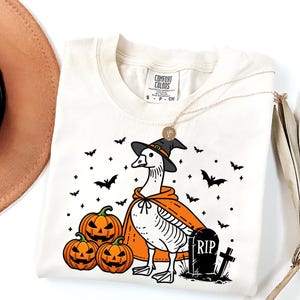 Puede incluir: Una camiseta blanca con una ilustraci&oacute;n en blanco y negro de un ganso con un sombrero de bruja y una capa. El ganso est&aacute; de pie junto a una l&aacute;pida que dice "RIP". Tambi&eacute;n hay calabazas y murci&eacute;lagos en la ilustraci&oacute;n.